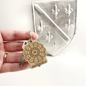 May include: A wooden keychain with a brown mandala design and the word "Srebrenica" engraved on the bottom. The keychain is being held by a hand.