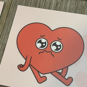 May include: A red heart-shaped sticker with a sad face and small arms and legs. The sticker has a pink background and a cartoonish design. The heart has a textured appearance and black outlines.