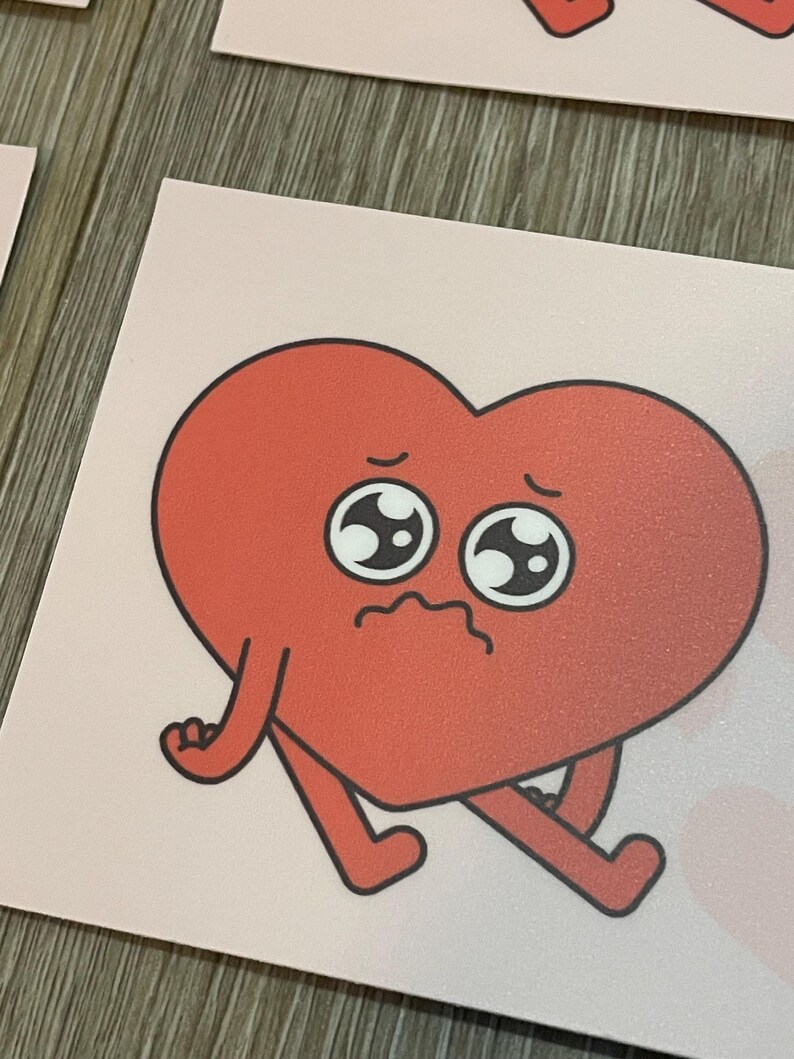 May include: A red heart-shaped sticker with a sad face and small arms and legs. The sticker has a pink background and a cartoonish design. The heart has a textured appearance and black outlines.