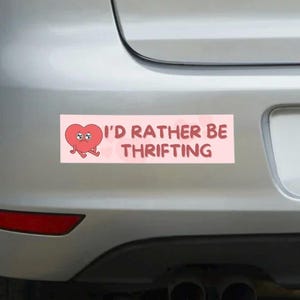 May include: A rectangular pink bumper sticker with the text "I'D RATHER BE THRIFTING" and a cartoon heart character. The heart is red with a sad expression and small arms and legs.