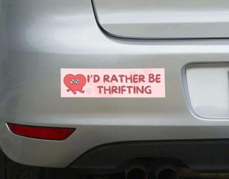 May include: A rectangular pink bumper sticker with the text "I'D RATHER BE THRIFTING" and a cartoon heart character. The heart is red with a sad expression and small arms and legs.