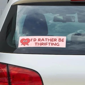 May include: A pink rectangular bumper sticker on a car's rear window. The sticker features a cartoon heart with eyes and legs, and the text "I'D RATHER BE THRIFTING" in white capital letters.