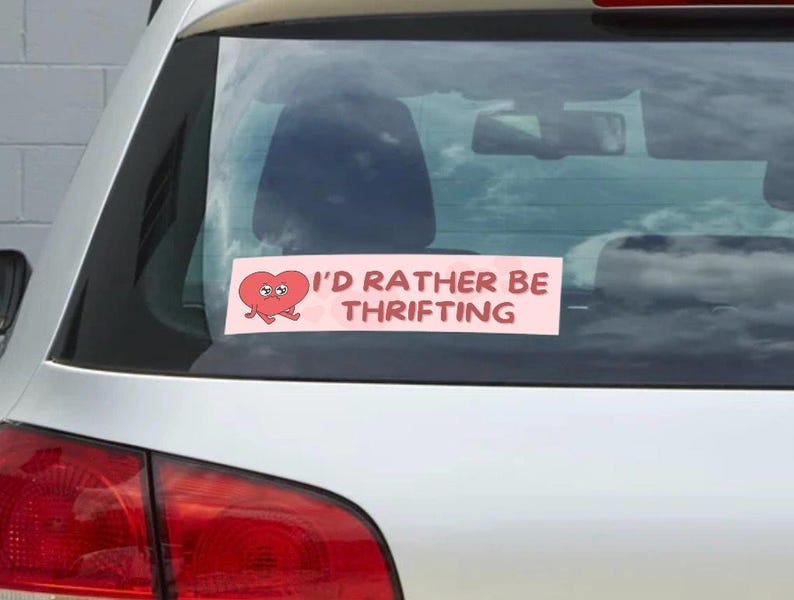May include: A pink rectangular bumper sticker on a car's rear window. The sticker features a cartoon heart with eyes and legs, and the text "I'D RATHER BE THRIFTING" in white capital letters.