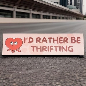 May include: A rectangular sign with the text "I'D RATHER BE THRIFTING" in brown letters. A red cartoon heart with sad eyes and legs is on the left side of the sign. The sign is on a gray surface.