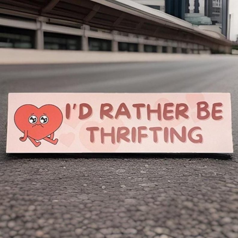 May include: A rectangular sign with the text "I'D RATHER BE THRIFTING" in brown letters. A red cartoon heart with sad eyes and legs is on the left side of the sign. The sign is on a gray surface.