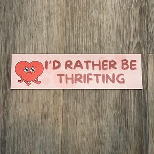 May include: A pink sticker with the text "I'D RATHER BE THRIFTING" in a dark pink font. The sticker features a cartoon heart with a sad expression and small arms and legs.