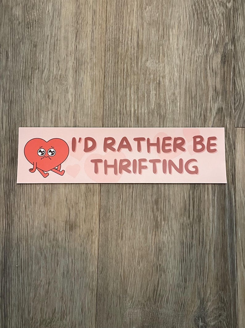 May include: A pink sticker with the text "I'D RATHER BE THRIFTING" in a dark pink font. The sticker features a cartoon heart with a sad expression and small arms and legs.