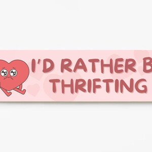 May include: A rectangular sign with a light pink background and the text "I'D RATHER BE THRIFTING" in a reddish-brown font. A cartoon heart with sad eyes and legs is on the left side.