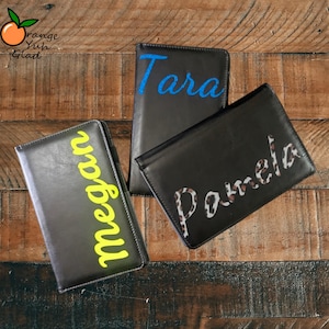 May include: Three black leather wallets with personalized names. One wallet has the name "Megan" in yellow, another has "Tara" in blue, and the third has "Pamela" in a leopard print design. The wallets are arranged on a wooden surface.