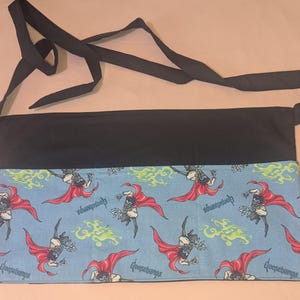 May include: A black and blue Goosebumps-themed apron with a long strap. The apron features a light blue lower section with a repeating graphic of a monster in a red cape, with the word "Goosebumps" printed below each graphic.