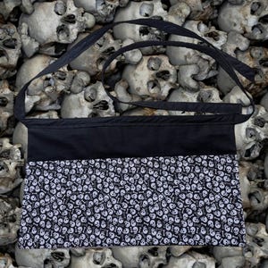 May include: Black apron with a black strap and a white skull and crossbones pattern on the bottom. The apron is designed to be worn around the waist and tied at the back.