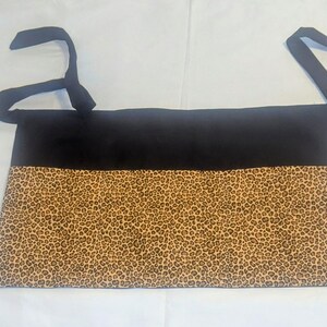 May include: A black apron with a leopard print bottom section and black ties. The apron is rectangular and has a simple design, suitable for kitchen or craft use. The leopard print is a tan color with dark brown spots.