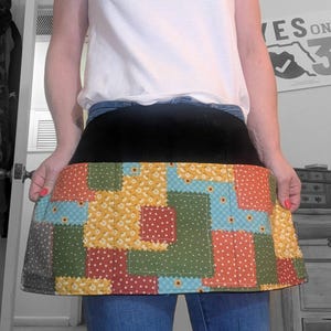 Peut inclure: Un tablier patchwork avec une ceinture noire et une jupe à motifs colorés. La jupe présente des carrés de tissu jaune, rouge, vert et bleu avec des motifs floraux et à pois. Le tablier est porté sur un jean bleu.