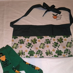 以下が含まれることがあります： 緑色のエプロンと、お揃いの緑色の靴下。エプロンは、クローバー模様で、濃い緑色のトップスと、クローバーと小さなラインストーン模様のボトムがあります。靴下は緑色で、レプラコーンのグラフィックと「Lucky and I Gnome It」の文字が入っています。