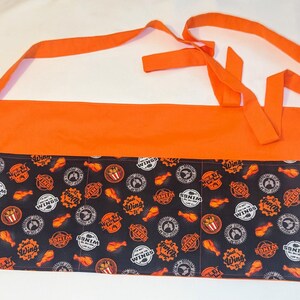 May include: Orange and black fabric apron with multiple pockets. The apron has a black and white print of chicken wings and the text "Just Wings".