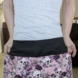 May include: A black and pink apron with a skull and floral pattern. The apron has a black waistband and a skirt with white skulls, purple flowers, and spiderwebs on a pink background.