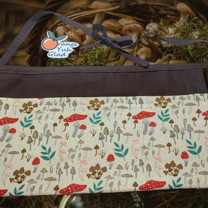 Puede incluir: Un delantal de lona marrón con estampado de setas y un bolsillo. El delantal tiene una correa marrón y una pegatina naranja y blanca que dice "Orange Yuh Glad".