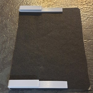 May include: A black, rectangular, felt-covered surface with two white plastic clips on the top and bottom edges.