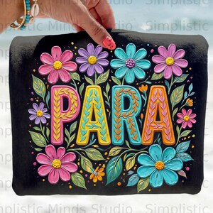 Könnte beinhalten: Schwarze Tragetasche mit dem Wort "PARA" in einem floralen Design. Das Design zeigt rosa, lila und türkisfarbene Blumen mit grünen Blättern, was eine lebendige Ästhetik ergibt.