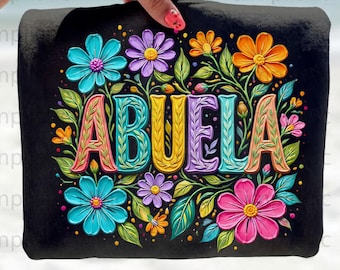 May include: Black tote bag featuring the word "ABUELA" in colorful, floral-style lettering. The design includes vibrant flowers in shades of blue, pink, orange, and purple, with green leaves. The bag is held by a hand.