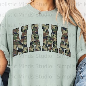 May include: A pale green t-shirt featuring the word "MAMA" in large, block letters with a camouflage design. The shirt has a loose fit and rolled sleeves. The text "Simplistic Minds Studio" is printed around the shirt.