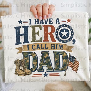 Può includere: Maglietta bianca con la scritta "I HAVE A HERO, I CALL HIM DAD." La parola "HERO" è in blu e rosso, e "DAD" è in mimetica. Include una bandiera americana e stivali militari.
