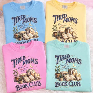 Op de afbeelding: Vier pastelkleurige crewneck sweatshirts: blauw, geel, roze en teal. Elk shirt toont de tekst "Tired Moms Book Club" met een afbeelding van boeken, planten, kaarsen en een halve maan. De shirts zijn gemaakt van een zacht materiaal.