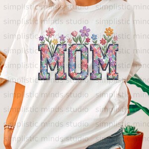 May include: White t-shirt with the word "MOM" in large, floral letters. The letters are decorated with colourful flowers. A casual, everyday wear item.
