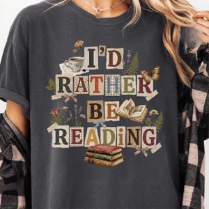 May include: Dark gray t-shirt with the text "I'd Rather Be Reading" in a collage style. The design includes a teacup, books, flowers, and a butterfly. The shirt is a comfortable, casual style.