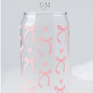 Puede incluir: Vaso transparente con forma de lata decorado con lazos y corazones rosas. El vaso tiene una superficie lisa y transparente y una abertura ancha. El diseño se repite en toda la superficie. En la esquina superior izquierda del vaso está el texto "SM" encima de "STUDIO".