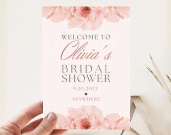 May include: A blush pink sign with floral accents, announcing "Welcome to Olivia's Bridal Shower" with the date 9.20.2023 and the word "ANYWHERE". The sign features watercolor-style pink flowers at the top and bottom.