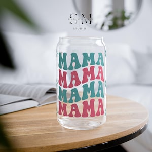 May include: A clear glass can-shaped drinking glass with the word "MAMA" repeated in teal and pink retro font. The glass sits on a wooden table. The top of the glass has a slight curve, and the glass is empty.
