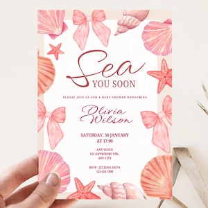 May include: Baby shower invitation with a watercolor design. The invitation features pink seashells, bows, and starfish. Text reads "Sea You Soon" and includes event details. The invitation is held by a hand.