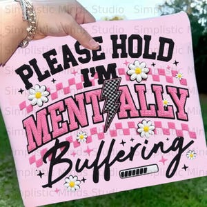 May include: A pink graphic with the text "PLEASE HOLD I'M MENTALLY Buffering" in black and pink. The design includes a checkered pattern, lightning bolt, daisies, and a loading bar.
