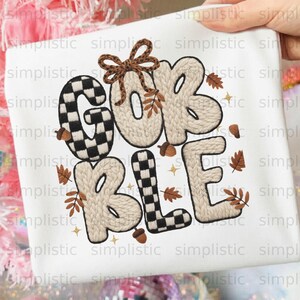 May include: A white t-shirt featuring the text GOBBLE in a mix of black and white checkered and beige cable knit patterns. The design includes a brown leopard print bow, small brown acorns, scattered autumn leaves, and tiny gold star accents.