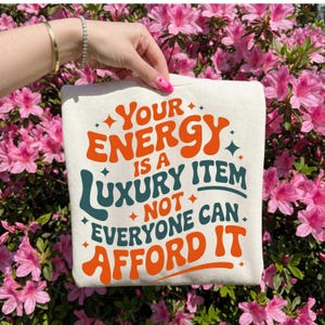 Puede incluir: Bolso tote color crema con el texto "Your Energy Is A Luxury Item Not Everyone Can Afford It" en letras naranjas y turquesas. El bolso se muestra sobre un fondo de flores rosas.
