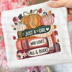 Op de afbeelding: Een wit t-shirt met een grafische print van een stapel van drie boeken met pompoenen en herfstbladeren erbovenop. De tekst op de ruggen van de boeken luidt JUST A GIRL WHO LOVES FALL & BOOKS. Een crèmekleurige sjaal ligt over de boekenstapel.