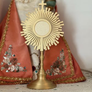May include: A gold-colored monstrance with a sunburst design, featuring a central circular space. The monstrance stands on a pedestal base. In the background, a statue of a religious figure is visible, draped in a red and gold garment.