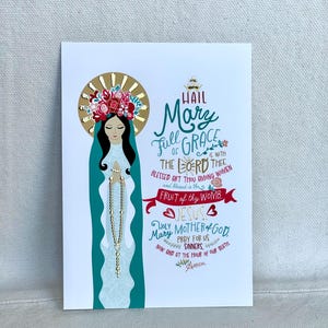 May include: A religious art print featuring an illustration of Mary with a gold halo and floral crown. The image includes the text "Hail Mary, Full of Grace" and other religious phrases in a decorative script. The artwork is on a white background.