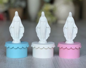 May include: Three white ceramic figurines of the Virgin Mary, each standing on a different colored base. The bases are light blue, white, and pink.