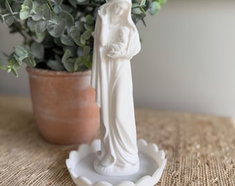 May include: White ceramic statue of the Virgin Mary holding a baby Jesus. The statue is on a white pedestal with a scalloped edge.