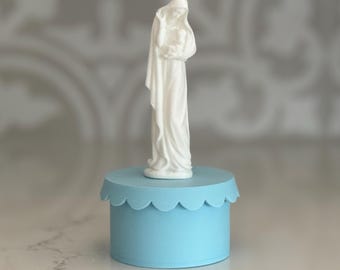 May include: White figurine of the Virgin Mary holding a baby Jesus, standing on a light blue pedestal with a scalloped edge.