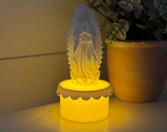 May include: White ceramic figurine of the Virgin Mary with a yellow glow emanating from the base. The figurine is on a white base with a scalloped edge.