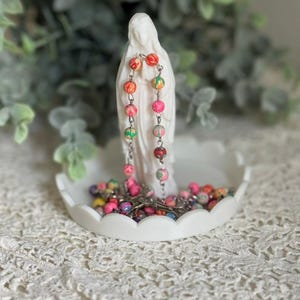 Peut inclure: Une figurine blanche repr&eacute;sentant une femme en longue robe est pos&eacute;e sur un plat blanc &agrave; bords festonn&eacute;s. Un chapelet aux perles multicolores &agrave; motifs roses, orange et verts est drap&eacute; sur la figurine, le reste des perles est enroul&eacute; dans le plat.