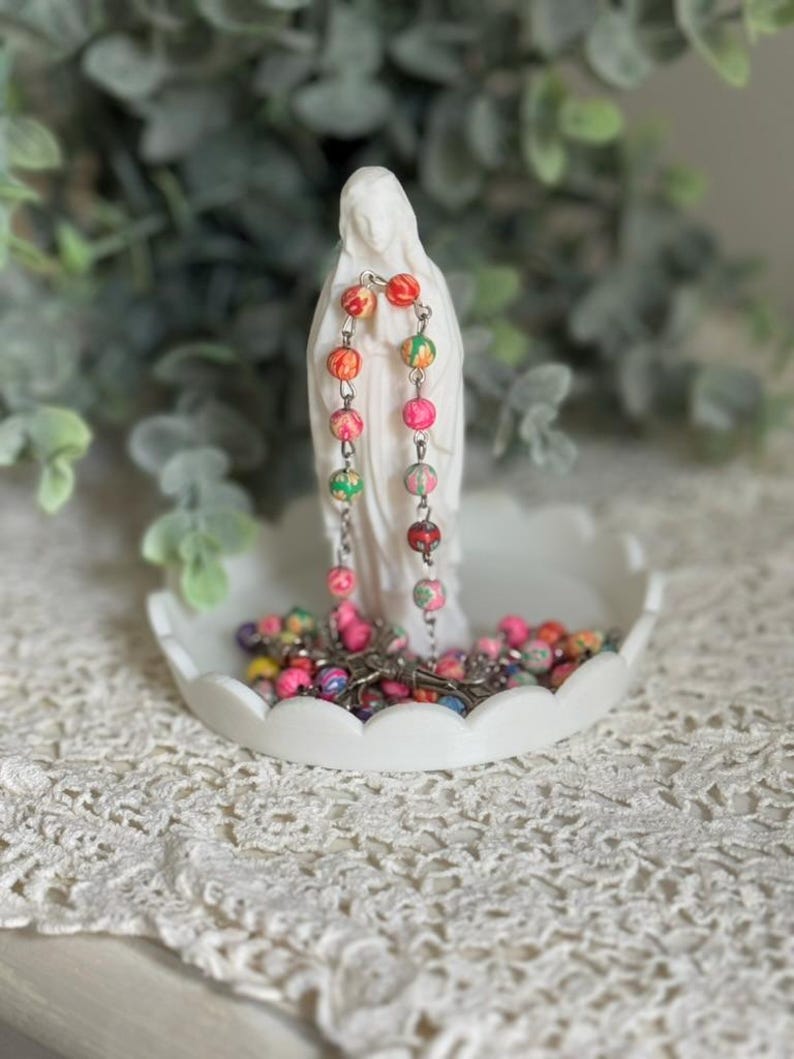 Peut inclure: Une figurine blanche repr&eacute;sentant une femme en longue robe est pos&eacute;e sur un plat blanc &agrave; bords festonn&eacute;s. Un chapelet aux perles multicolores &agrave; motifs roses, orange et verts est drap&eacute; sur la figurine, le reste des perles est enroul&eacute; dans le plat.