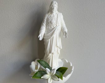 May include: A white religious statue of Jesus with outstretched arms, standing above a white, shell-shaped basin with white flowers and green leaves. The statue is set against a plain, light gray wall.