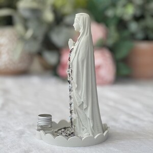 Peut inclure: Une figurine blanc mat repr&eacute;sentant une silhouette en robe &agrave; capuche, les mains jointes en pri&egrave;re, pos&eacute;e sur une base circulaire festonn&eacute;e. Un chapelet de perles claires et sombres pend des mains, et un petit flacon en verre &agrave; bouchon argent&eacute; est pos&eacute; sur la base.