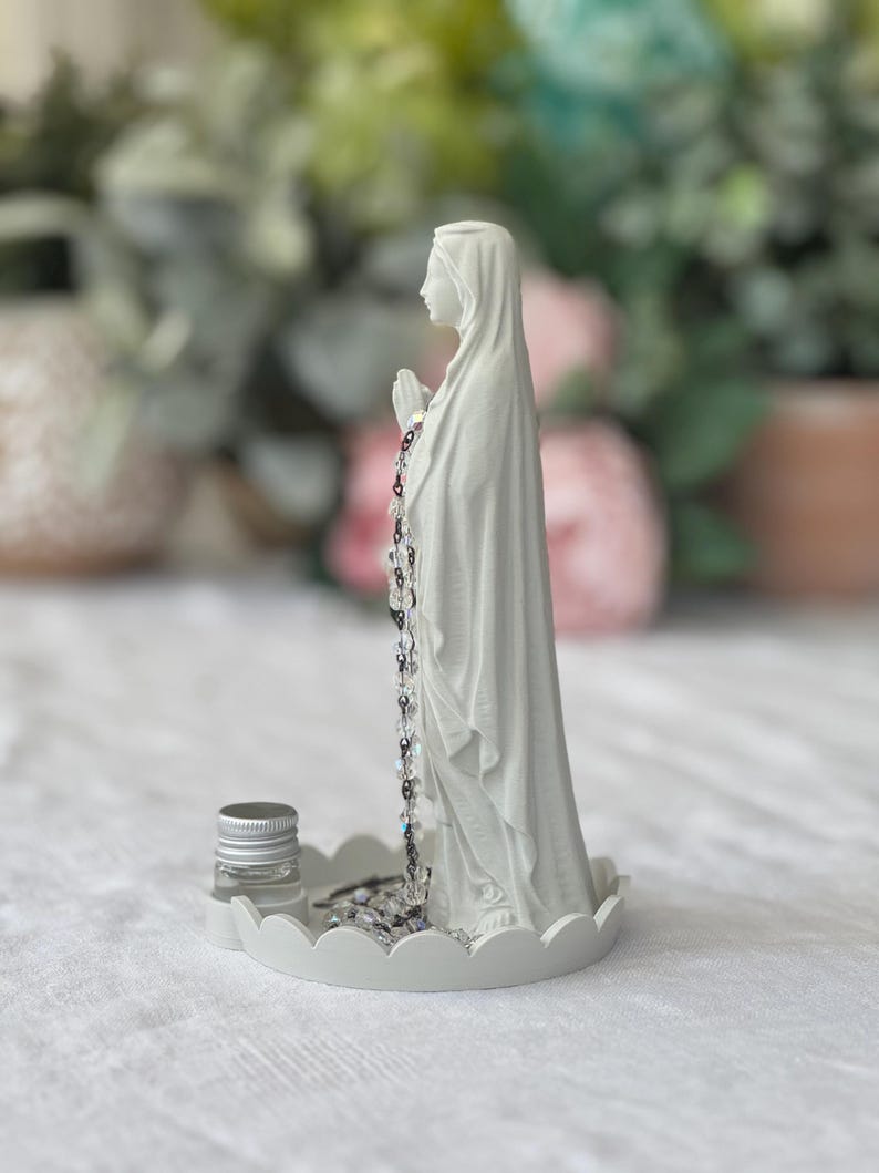 Peut inclure: Une figurine blanc mat repr&eacute;sentant une silhouette en robe &agrave; capuche, les mains jointes en pri&egrave;re, pos&eacute;e sur une base circulaire festonn&eacute;e. Un chapelet de perles claires et sombres pend des mains, et un petit flacon en verre &agrave; bouchon argent&eacute; est pos&eacute; sur la base.