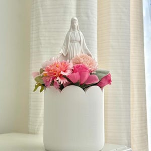 May include: A white cylindrical planter with a scalloped top edge containing an arrangement of artificial pink flowers and green leaves, featuring a small white figurine of the Virgin Mary standing centrally among the floral display.