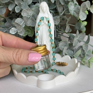 Peut inclure: Un petit flacon en verre transparent avec un bouchon &agrave; vis dor&eacute;, un collier de perles bleu clair &agrave; facettes avec des entretoises dor&eacute;es, et une figurine blanche repr&eacute;sentant une personne en pri&egrave;re, sur un plateau blanc festonn&eacute;.